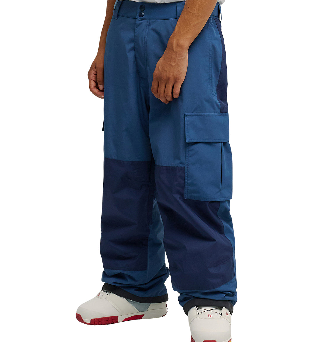 A person is wearing DC Shoes’ Mountain Lab Pants 10K—loose-fitting, breathable cargo pants in two shades of blue—paired with white sneakers featuring red accents. Only the pants and lower legs are visible against a plain white background.
