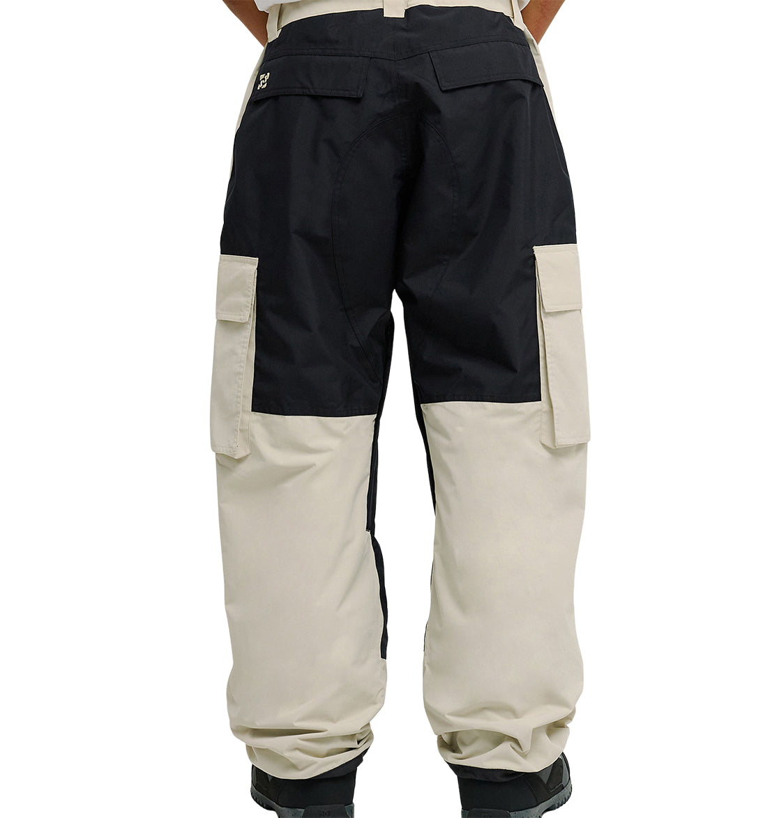 Wearing DC Shoes’ Mountain Lab Pants 10K, which showcase a two-tone design with black on the upper section and beige on the lower section, a person stands with their back to the camera. These pants deliver both standout style and snow-ready performance, featuring large side pockets, while black boots are partially visible.