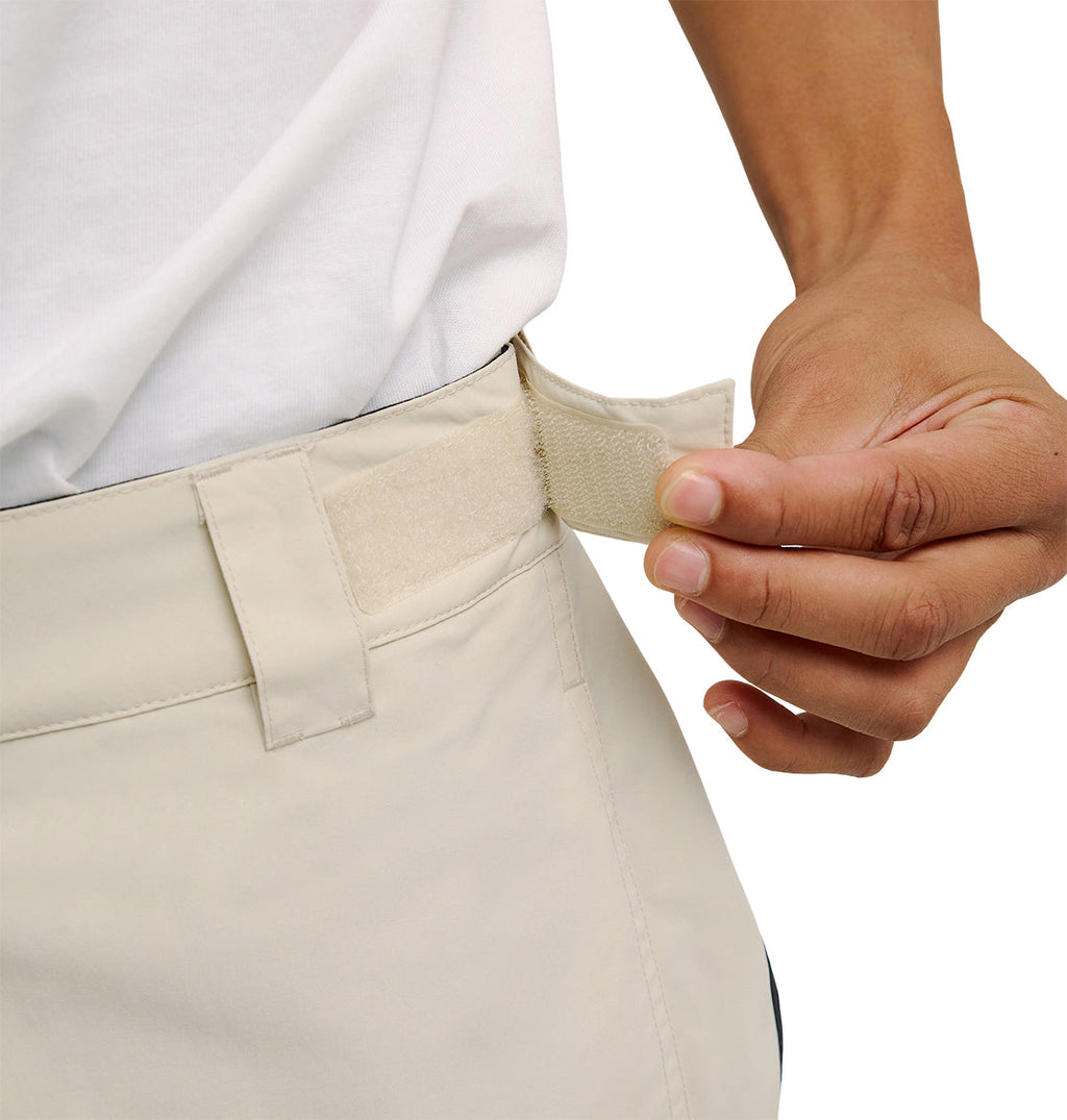 Wearing a white shirt, a person adjusts the hook-and-loop (Velcro) waistband strap of light-colored DC Shoes Mountain Lab Pants 10K, ensuring a comfortable fit and optimal performance in the snow.