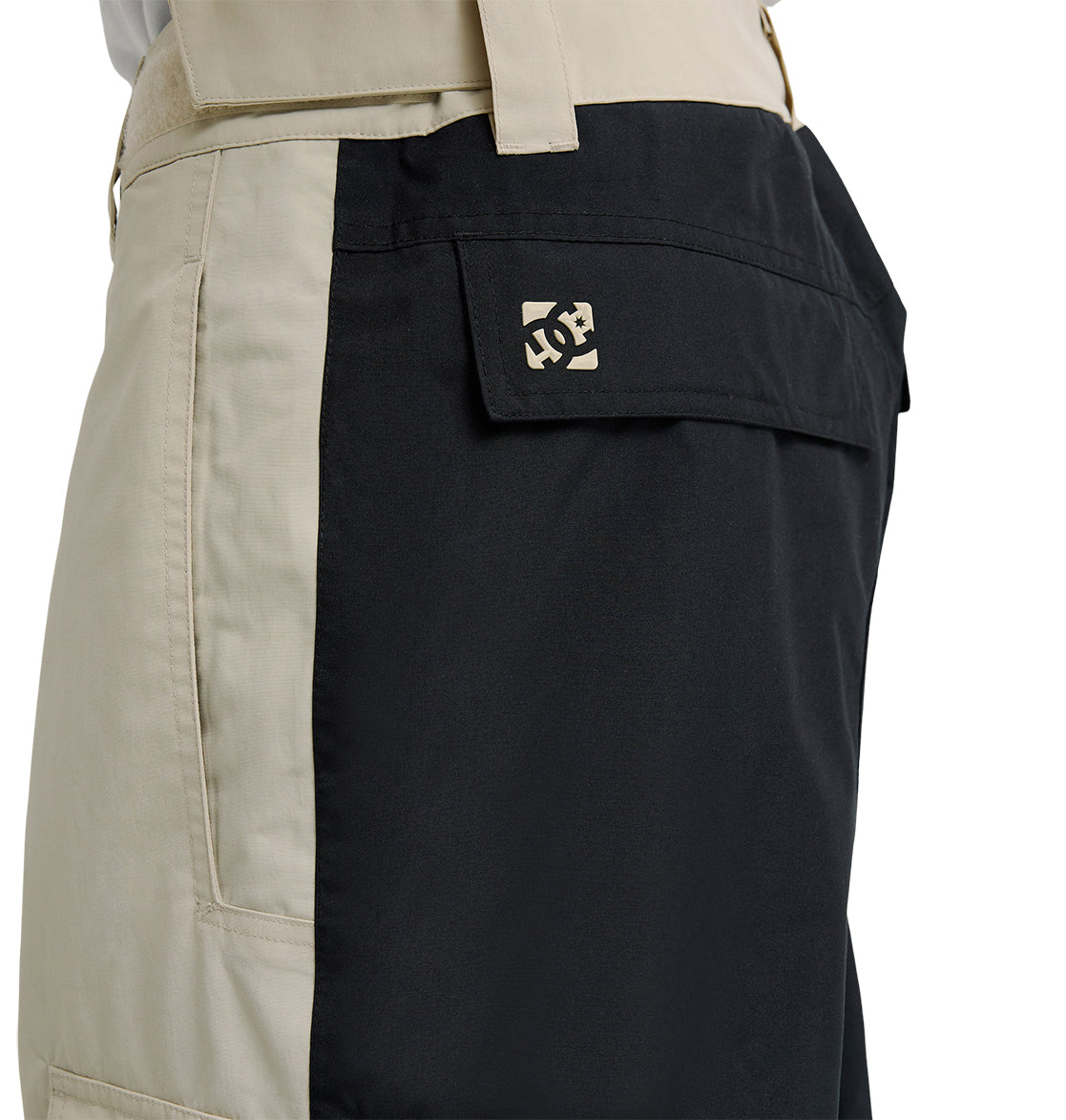 Side view of the DC Shoes Mountain Lab Pants 10K featuring beige and black color-blocked panels, with a flap pocket on the black section accented by a small, light-colored logo. Visible stitching and belt loops enhance the design, while breathable protection ensures comfort during active wear.
