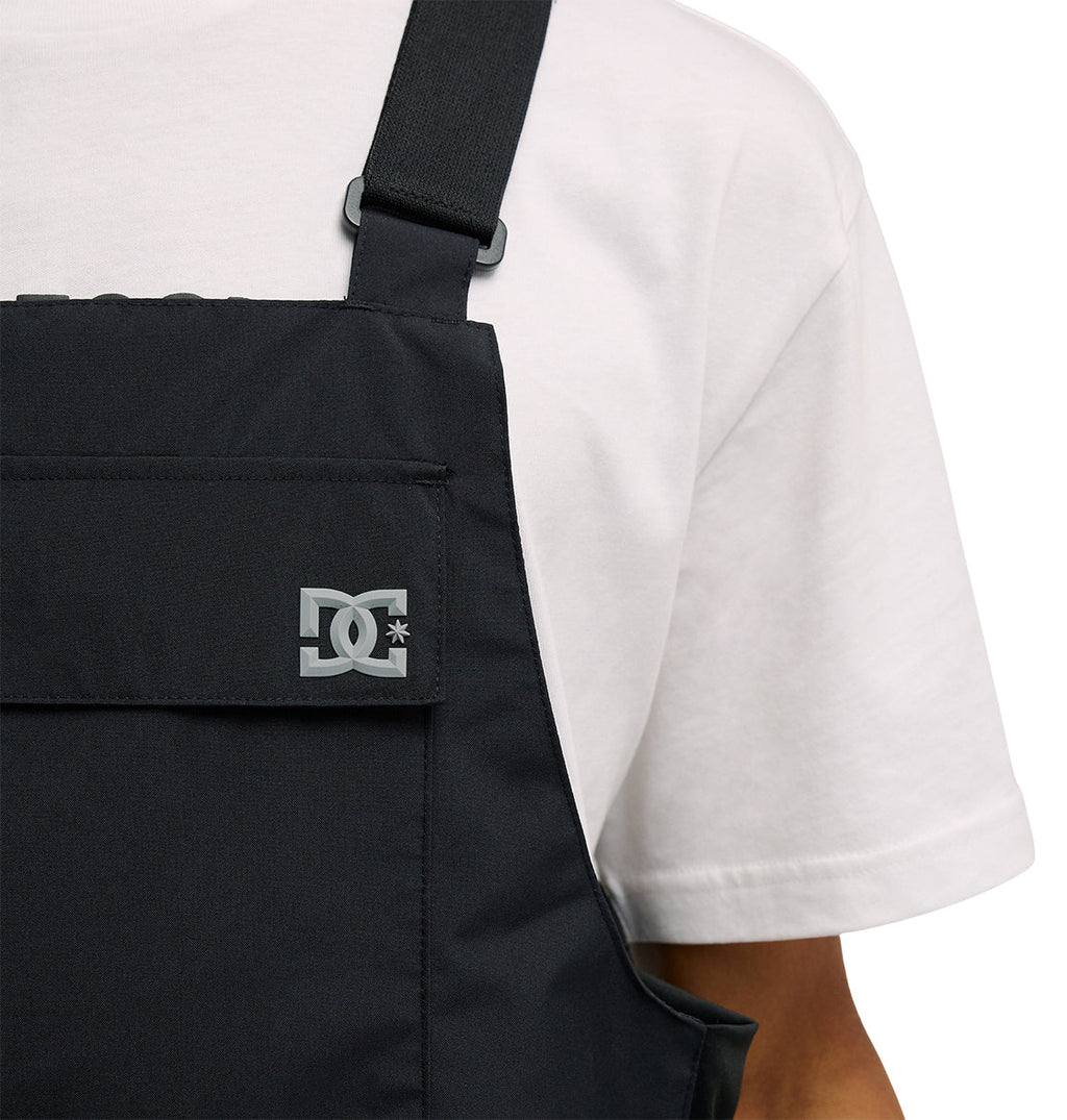 A person wearing the Docile Bib 10K by DC Shoes—black insulated overalls featuring the DC Shoes logo on the chest—layered over a white t-shirt, shown against a plain white background.