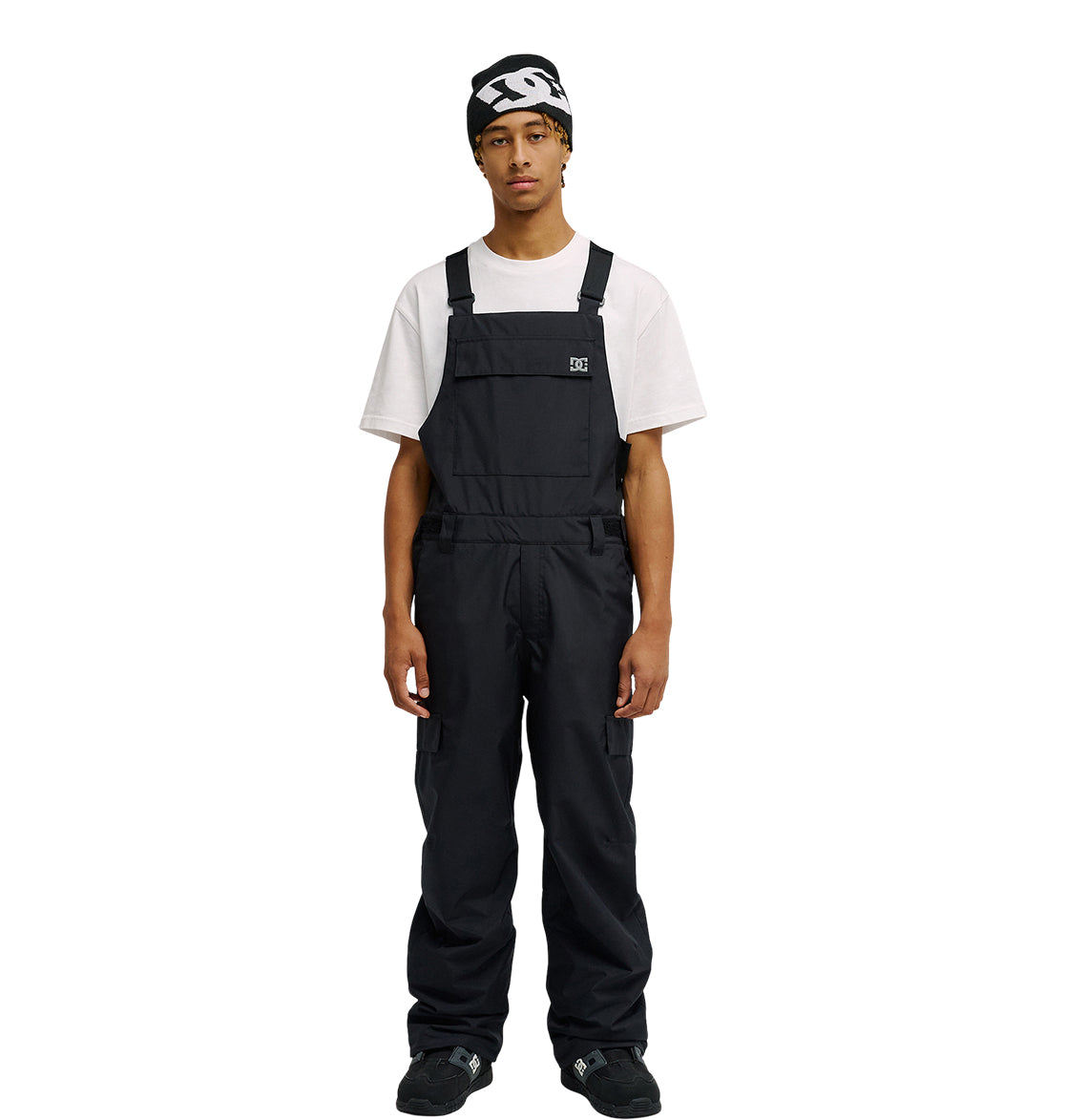 A person stands facing forward against a plain white background, wearing the DC Shoes Docile Bib 10K black snow bib overalls over a white T-shirt, paired with black shoes and a black beanie featuring a white logo.