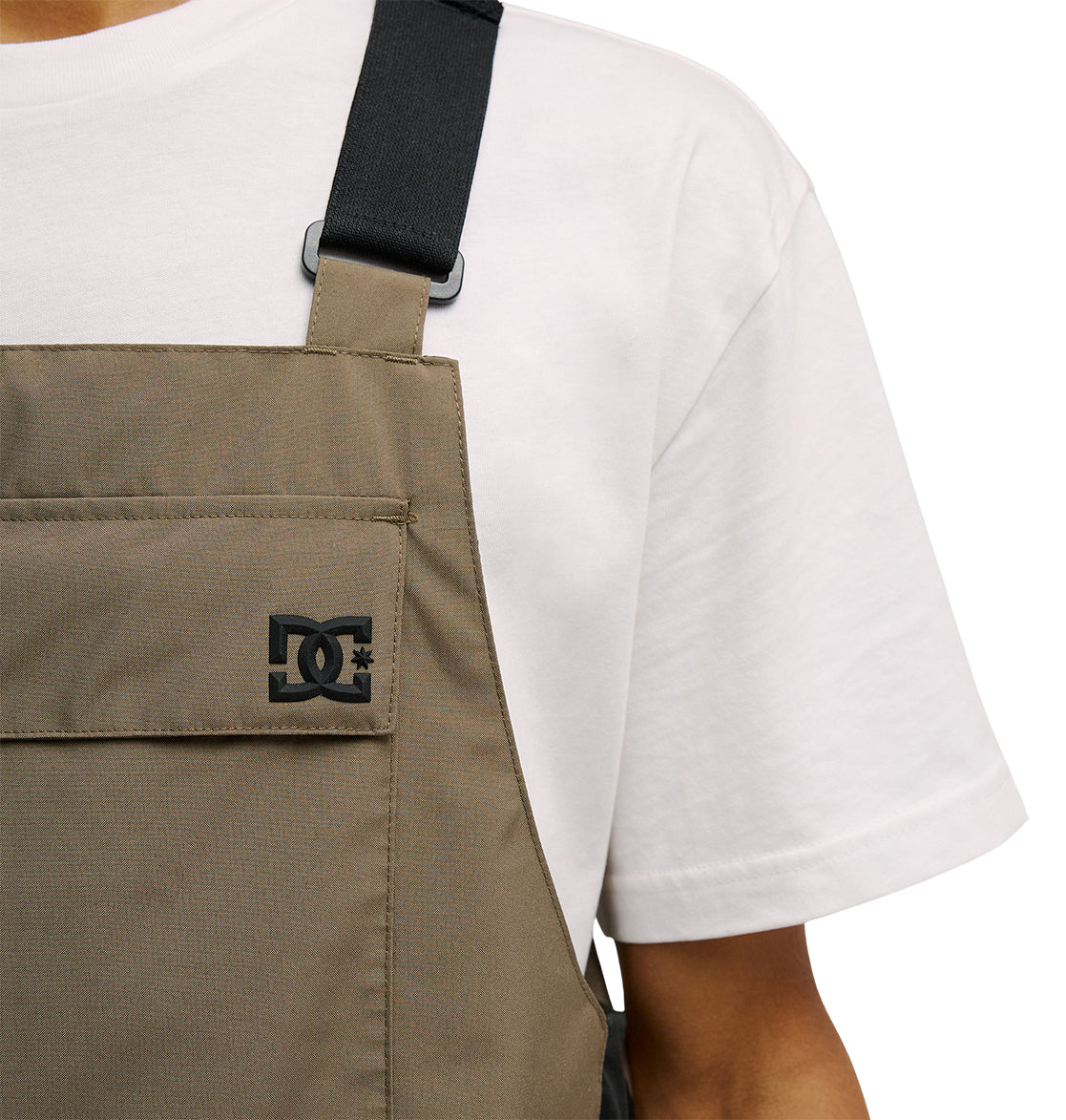 A person wearing the DC Shoes Docile Bib 10K in brown over white, featuring a black DC logo on the chest pocket and designed with breathable protection for added comfort; only the torso and part of one arm are visible.