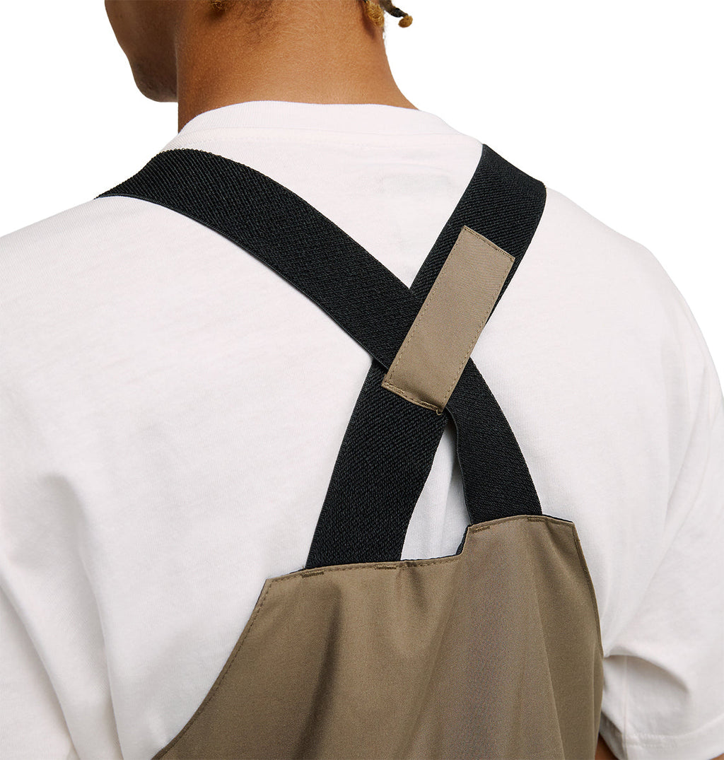 A close-up shows a person wearing the DC Shoes Docile Bib 10K—a white shirt paired with the brown apron featuring black straps that cross at the back. The image focuses on the upper back and shoulders, highlighting the apron’s breathable protection, with the wearer’s face out of view.