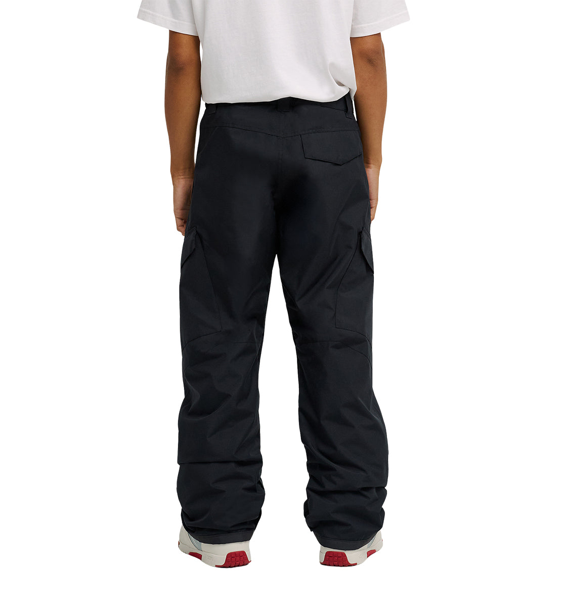 A person is shown from the waist down, wearing a white t-shirt and DC Shoes Banshee Pants—black cargo pants with waterproof insulation—paired with gray and red sneakers. Their back faces the camera.