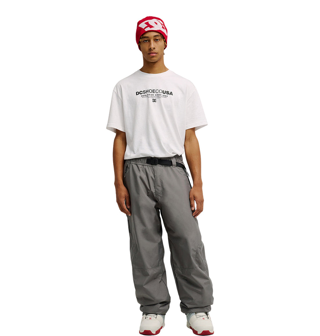 A young person wearing a red beanie, white graphic T-shirt, DC Shoes Smoke Pants 10K in gray with a black belt, and white sneakers stands facing the camera against a plain white background.