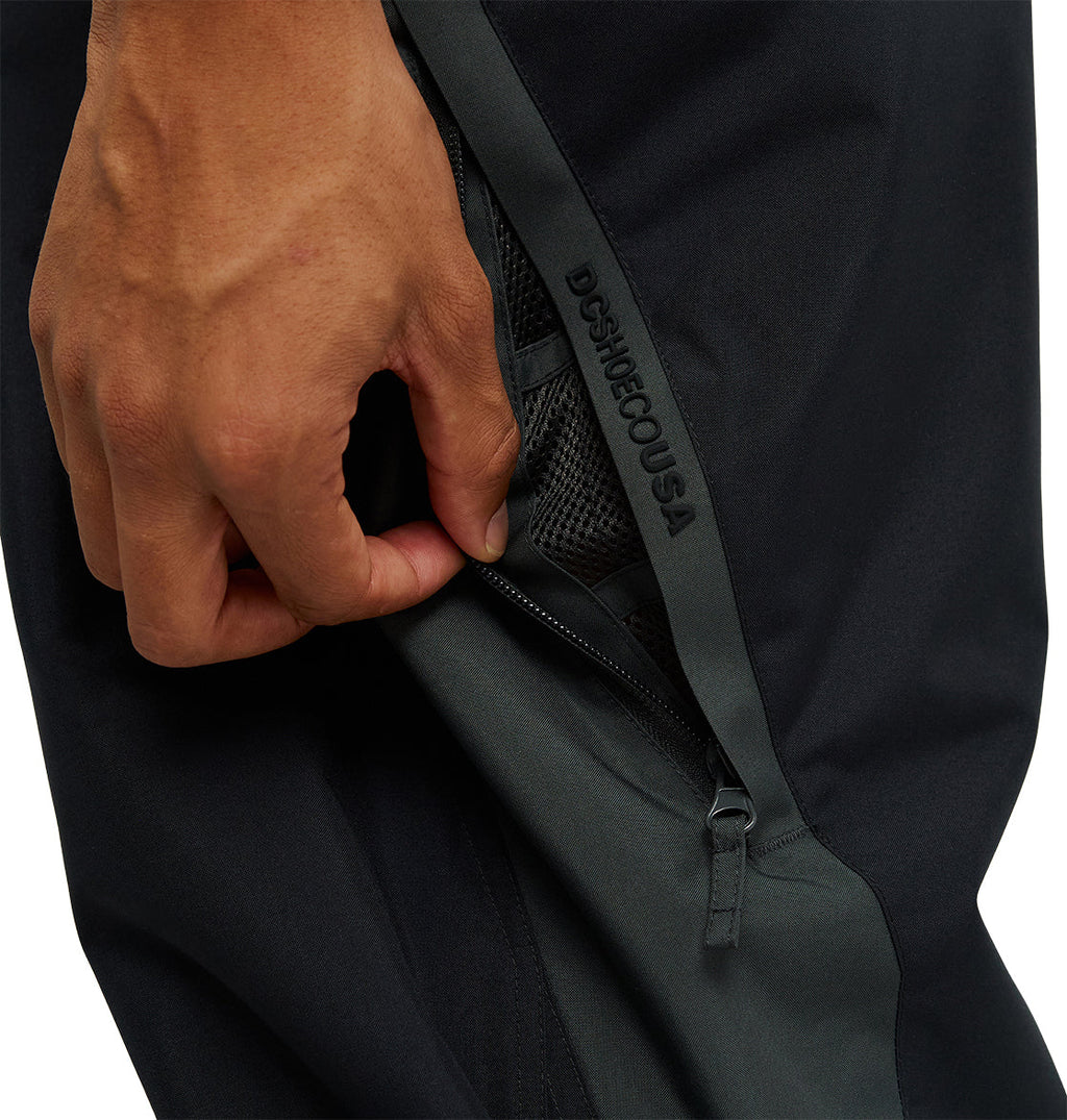 A hand pulls down the zipper of a black and dark gray mesh pocket on the DC Shoes Smoke Pants 10K, offering breathable protection for active days outdoors. The DC Shoes branding is visible along the zipper edge.