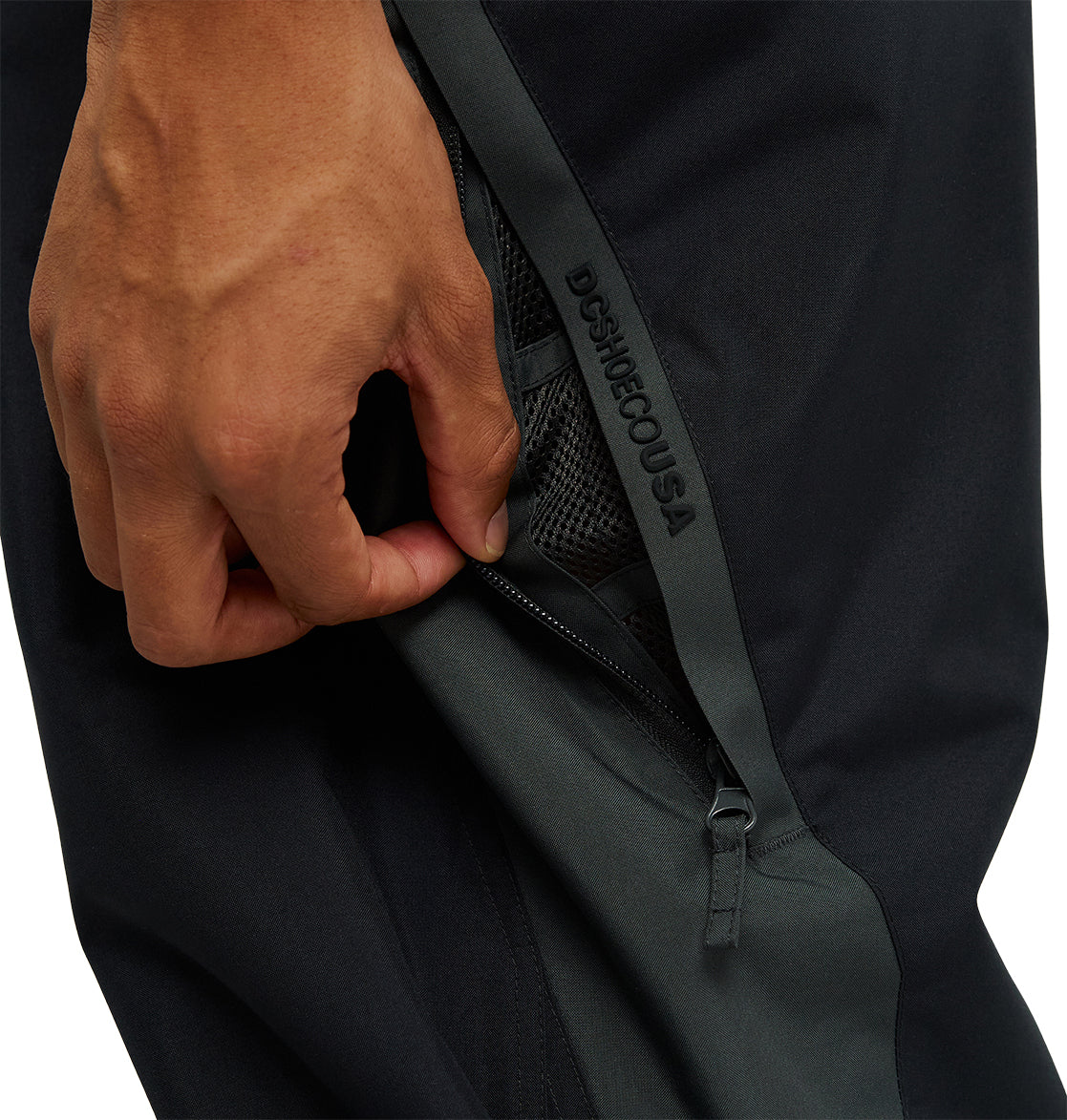 A hand pulls down the zipper of a black and dark gray mesh pocket on the DC Shoes Smoke Pants 10K, offering breathable protection for active days outdoors. The DC Shoes branding is visible along the zipper edge.