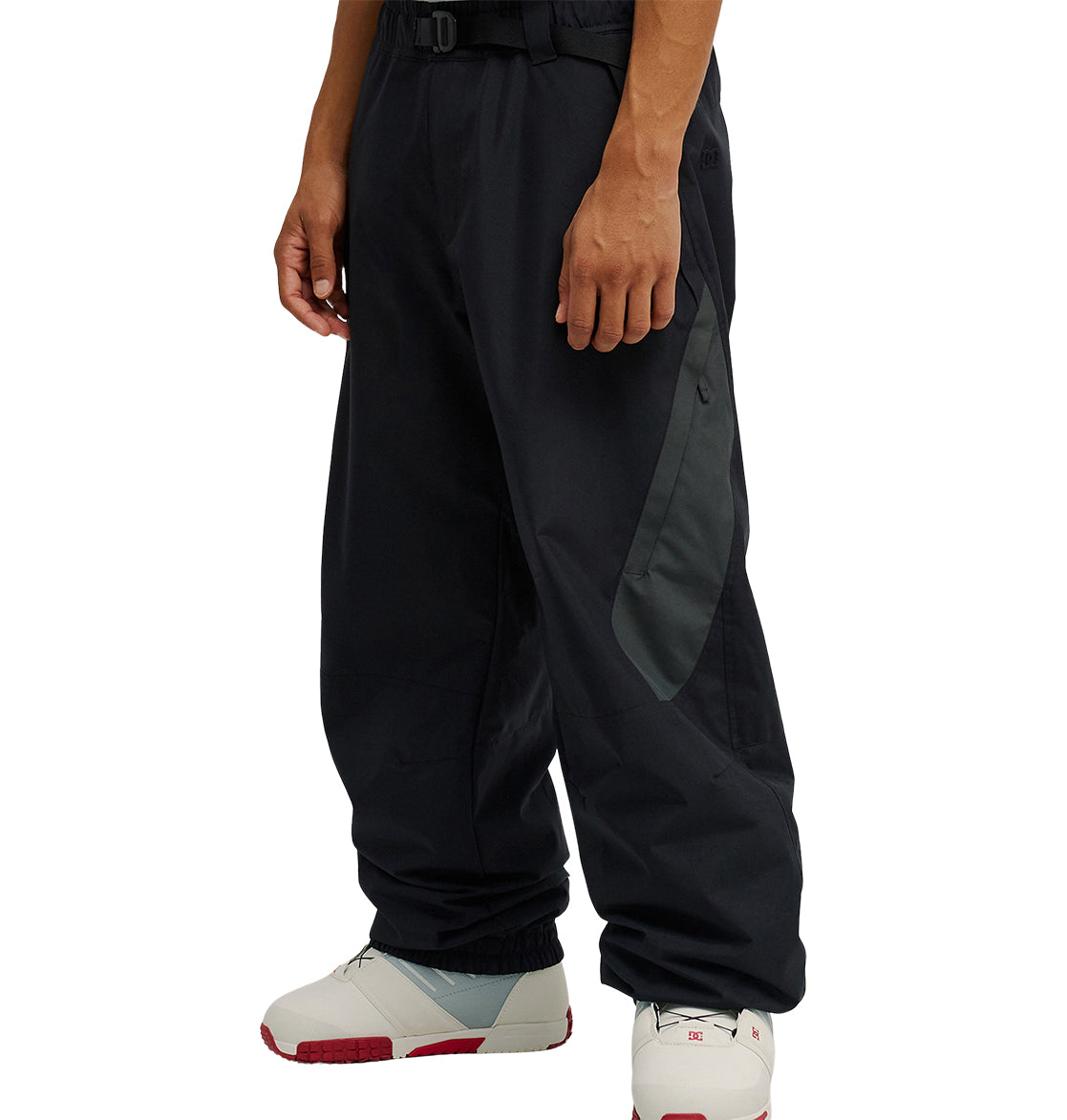 A person is wearing the DC Shoes Smoke Pants 10K in black and dark gray, featuring a side pocket and a belted waist, paired with white sneakers with red soles, while standing against a plain white background. The Smoke Pants 10K offer breathable protection and are engineered by DC Shoes for excellent snow performance, combining style with functionality.