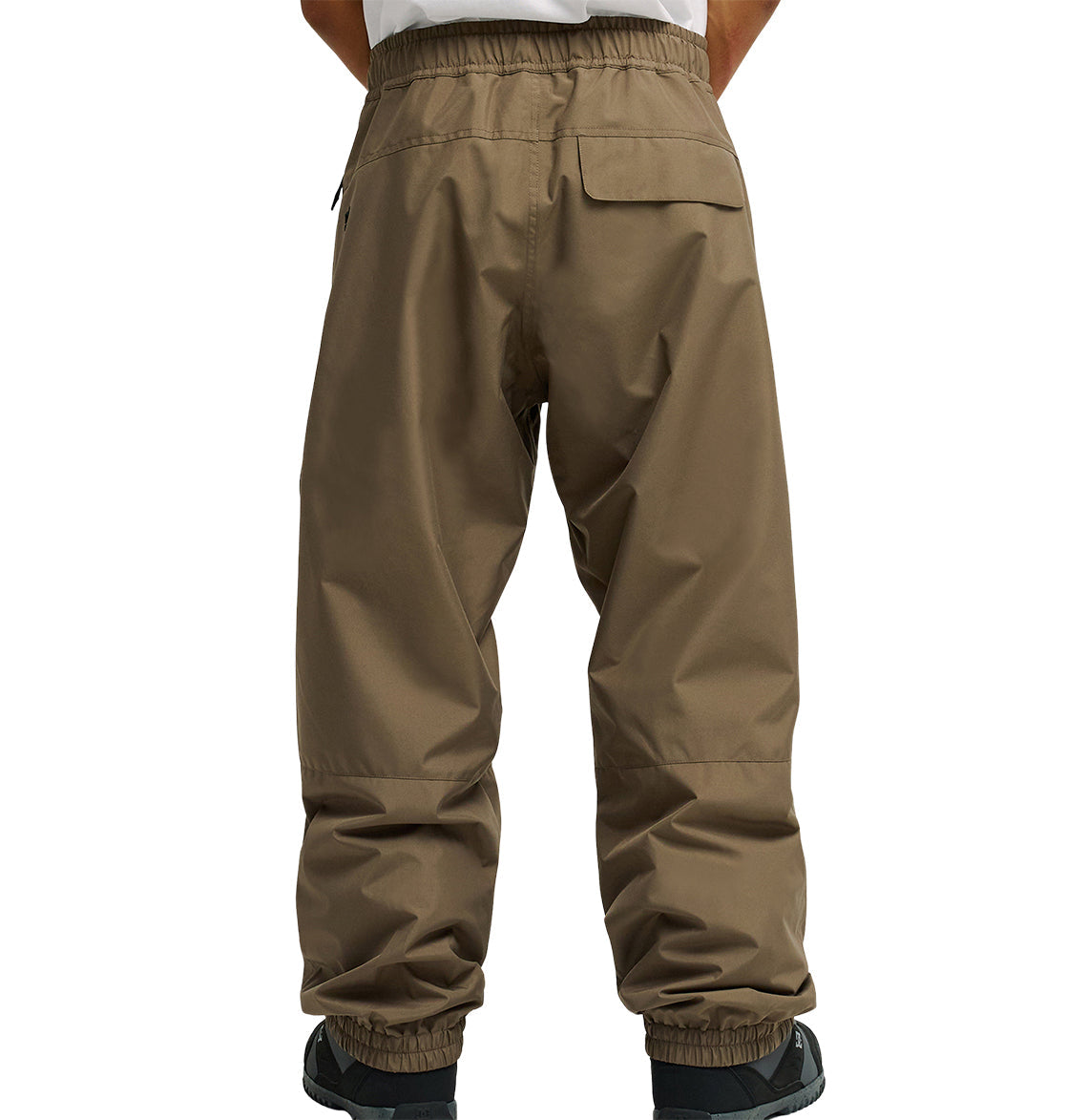 A person is seen from behind wearing DC Shoes Smoke Pants 10K—loose-fitting brown pants with an elastic waistband and cuffs. These breathable pants feature a flap back pocket, and only the lower back and legs are visible.