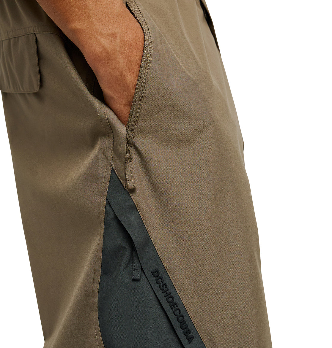 A close-up of a person’s hand in the pocket of DC Shoes Smoke Pants 10K snow pants in olive green, featuring a zipper and a black strip labeled DCSHOECOUSA, highlights their breathable and waterproof design.