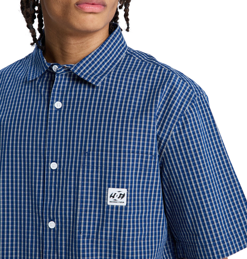 A person wearing the Reebok Game On Ss blue and white checkered short-sleeve button-up shirt with a white chest pocket label. Their face is partially visible.