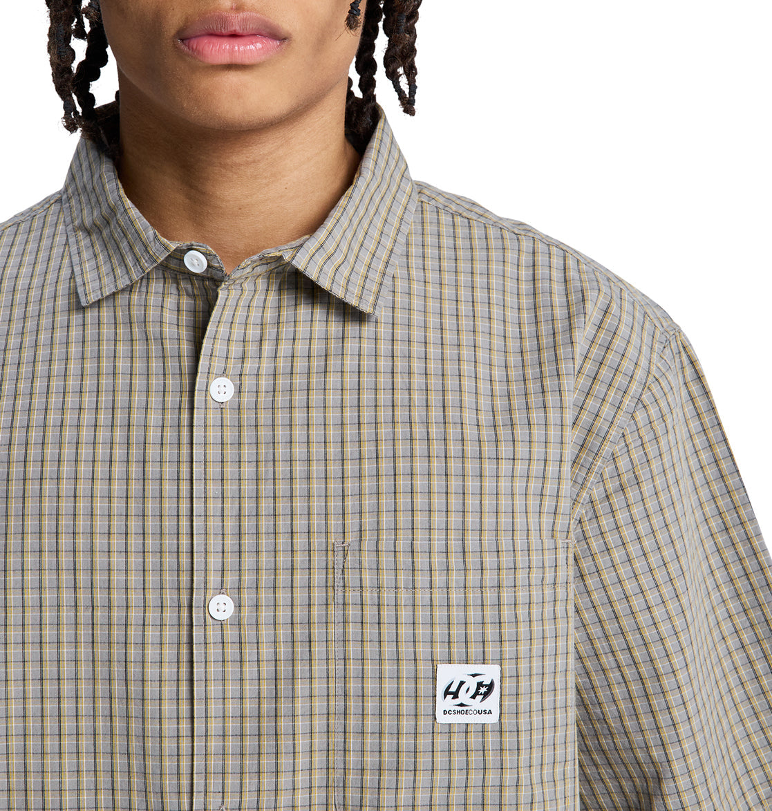 A person wears a beige and blue plaid Reebok Game On Ss button-up shirt with a chest pocket and rectangular logo patch. Only their lower face and upper torso are visible.