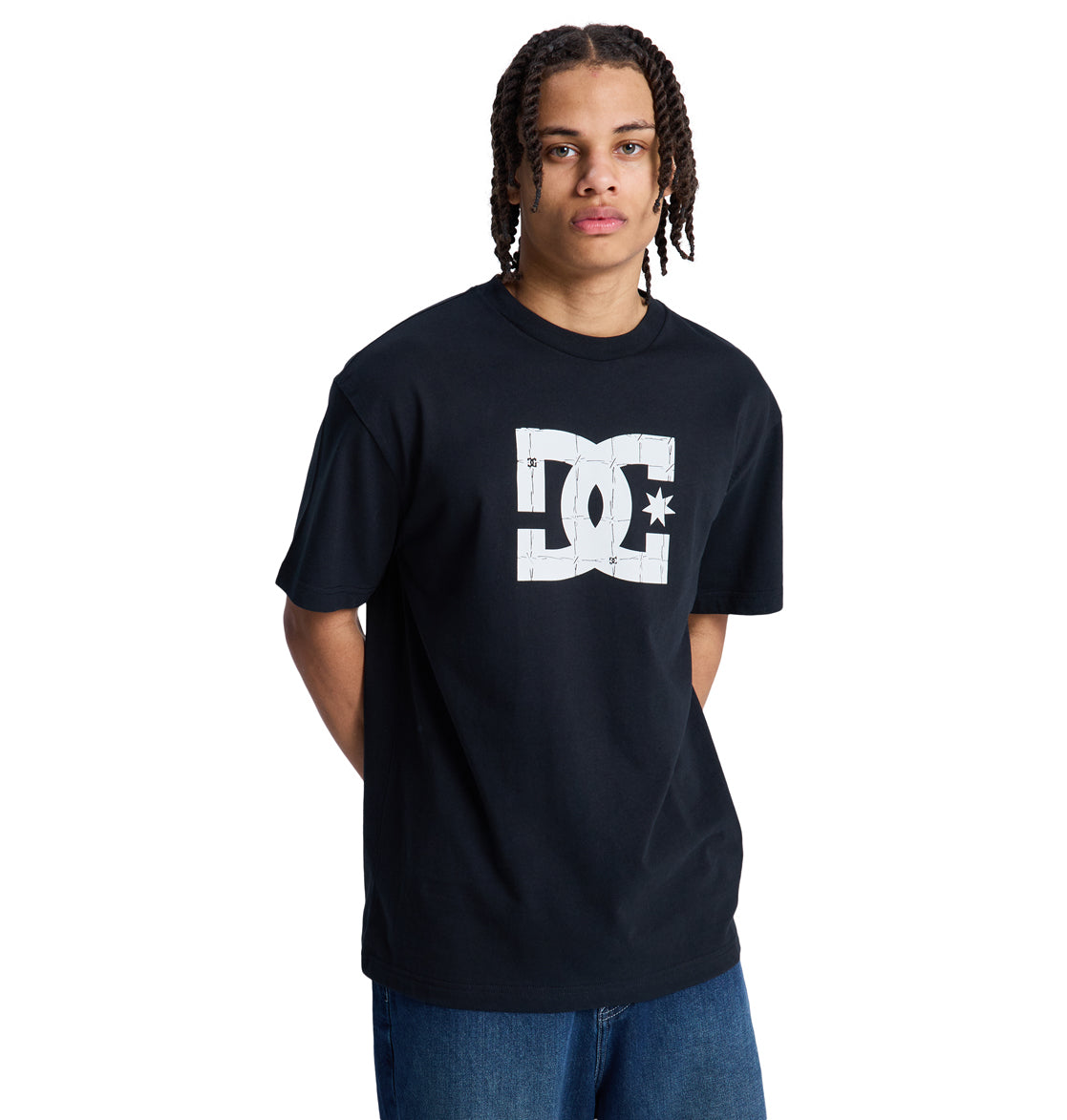A person with medium-length braids wears a black Reebok DC Star Filled Hss t-shirt with a large white logo on the front and blue jeans, standing against a plain white background.