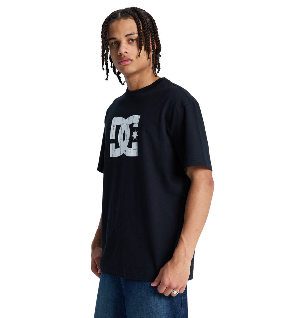 A person with braided hair wears a black Reebok DC Star Filled Hss t-shirt and blue jeans, standing against a white background.