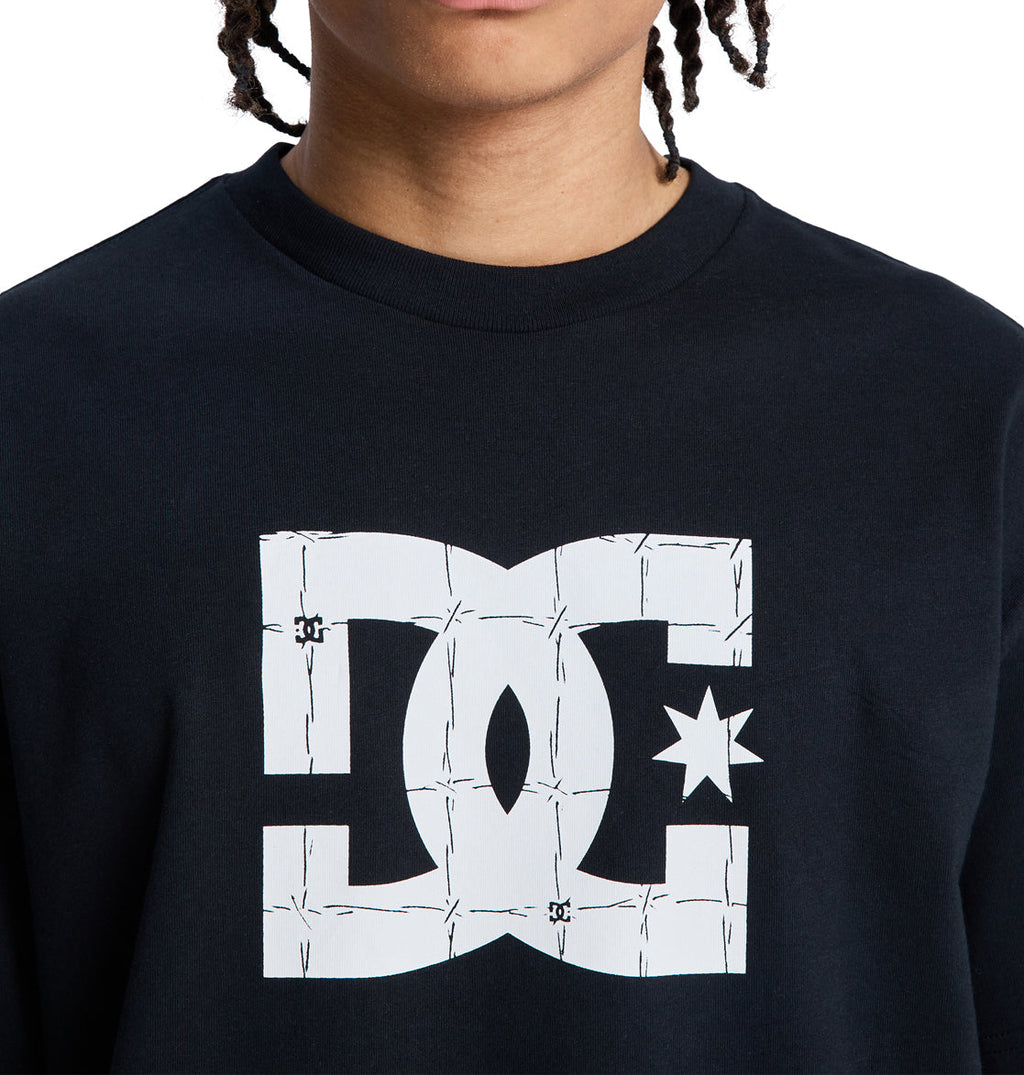 A person wears a black Reebok DC Star Filled Hss shirt with a large white logo on the front featuring a barbed wire design. Only the lower half of their face is visible.