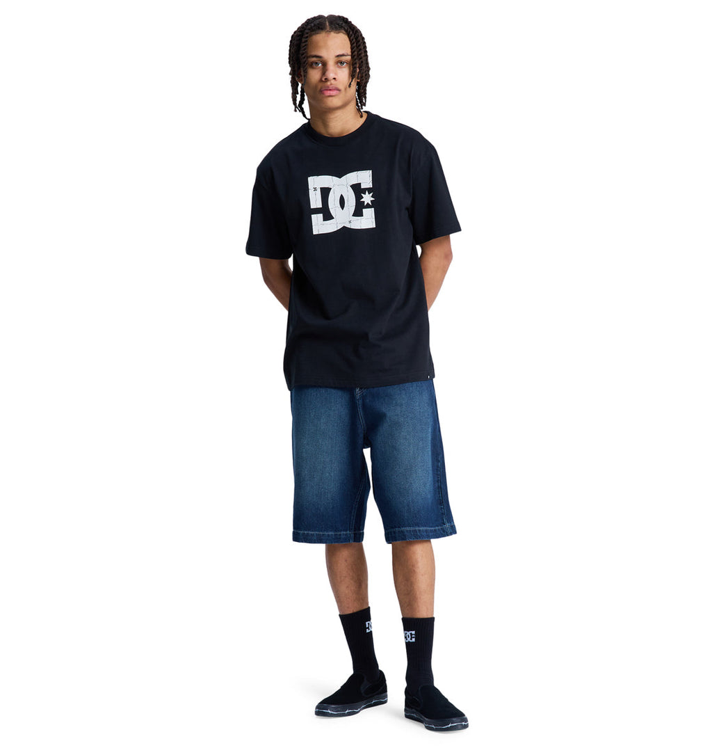 A young person with braided hair wears a black Reebok DC Star Filled Hss t-shirt, blue knee-length denim shorts, black DC socks, and black slip-on shoes, standing against a plain white background.