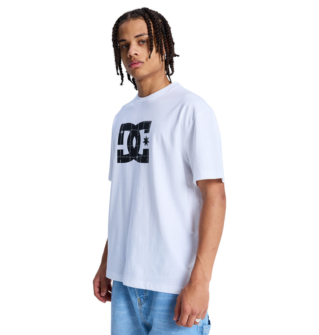 A young man with braided hair wears the Reebok DC Star Filled Hss, a loose white T-shirt with a bold black DC logo on the chest, paired with light blue jeans, standing against a plain white background.