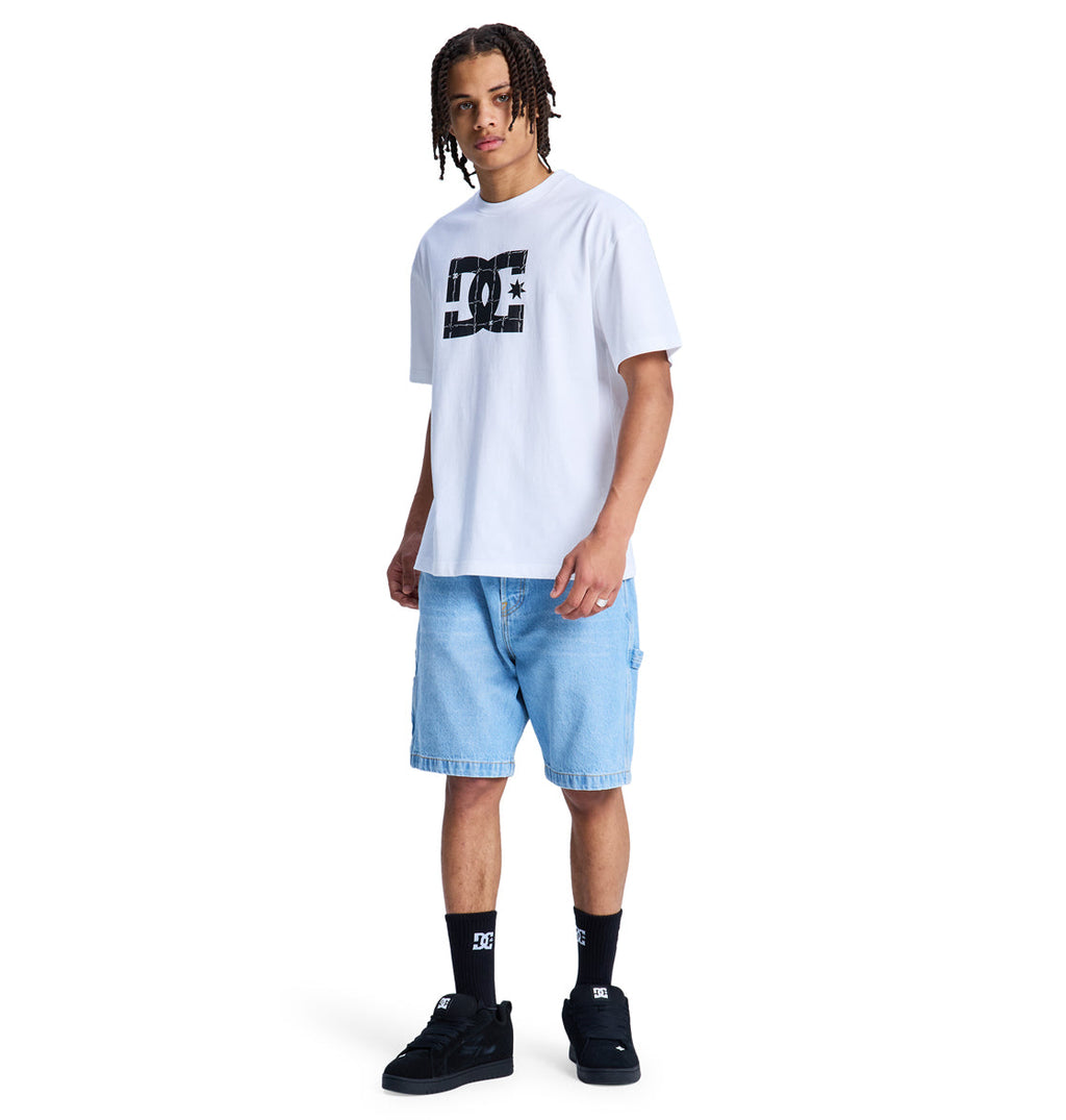 A young man with short dreadlocks stands against a plain white background, wearing a Reebok DC Star Filled Hss white t-shirt, light blue denim shorts, black DC logo socks, and black sneakers.