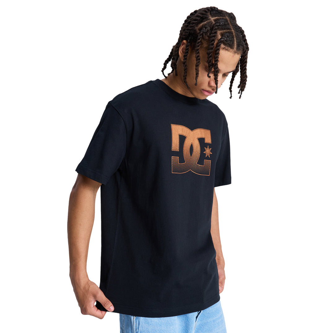 A person with braided hair wears a black Reebok DC Star Oxidized Hss t-shirt featuring a large bronze DC logo on the chest and light blue jeans, looking down against a plain white background.