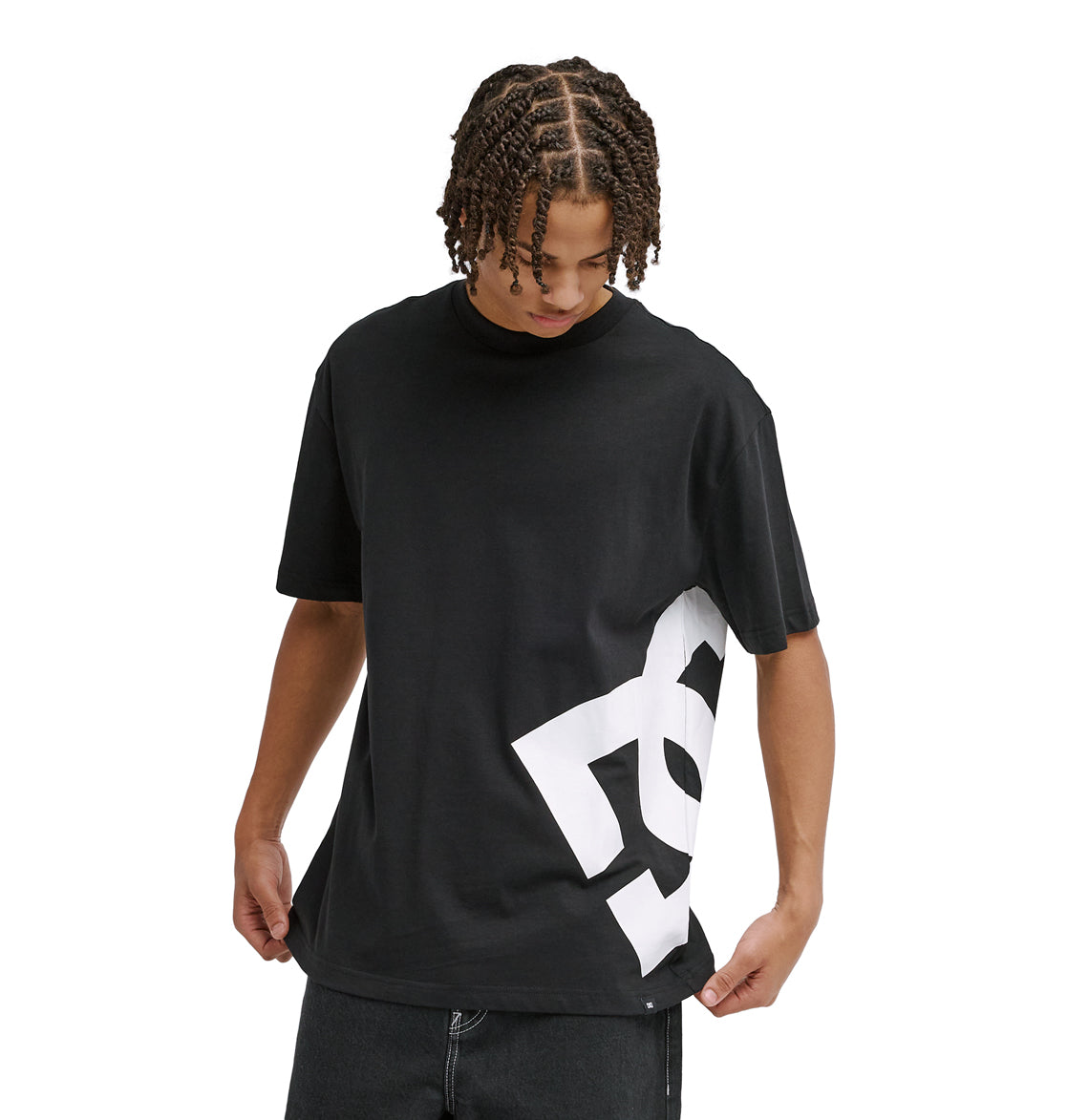 A person with short, braided hair looks down and holds the hem of a black Reebok DC Lanai Hss t-shirt featuring a large white graphic on the lower side.