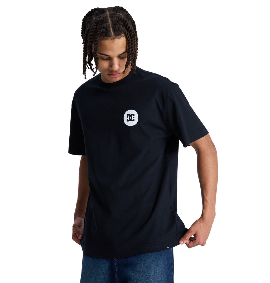 A person with braided hair looks down, wearing a loose-fitting black Reebok Shape Or Destroy Hss t-shirt and blue jeans, standing against a plain white background.