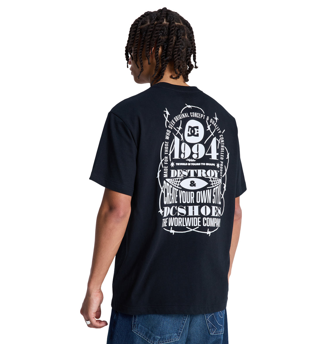 A person with short, twisted hair faces away from the camera wearing a Reebok Shape Or Destroy Hss black T-shirt, featuring a large white graphic with bold text, numbers, and barbed wire designs on the back.