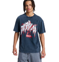 A person with braided hair wears a loose-fitting, dark blue DC Death Machine Hss graphic T-shirt by Reebok with white and red lettering and designs, paired with light blue shorts, standing against a plain white background.