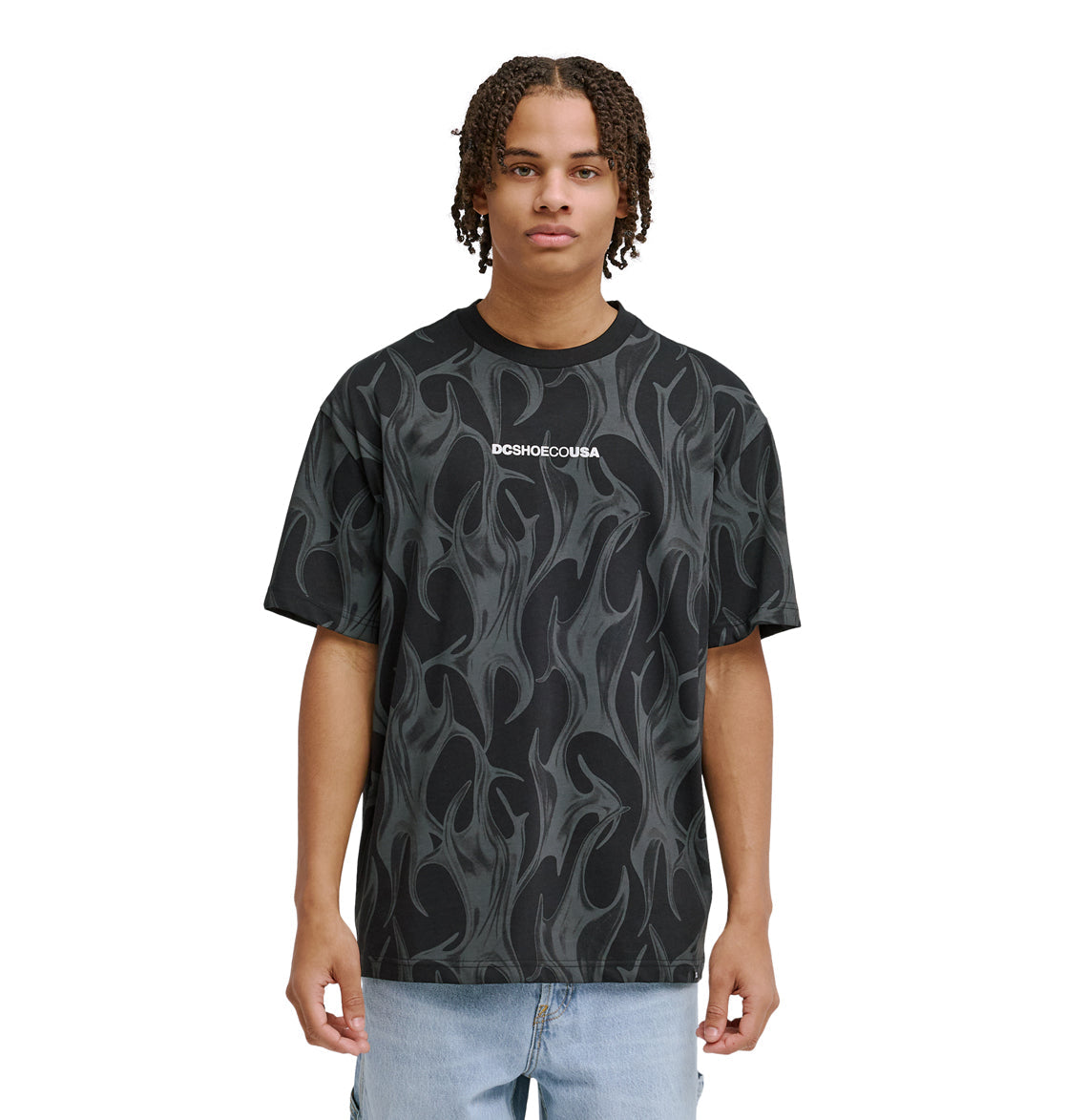 A young person with short, curly hair wears the Reebok DC Liquid Fuego Hss t-shirt—black with a gray flame pattern and "Reebok" printed in white on the chest—paired with light blue jeans, standing against a plain white background.
