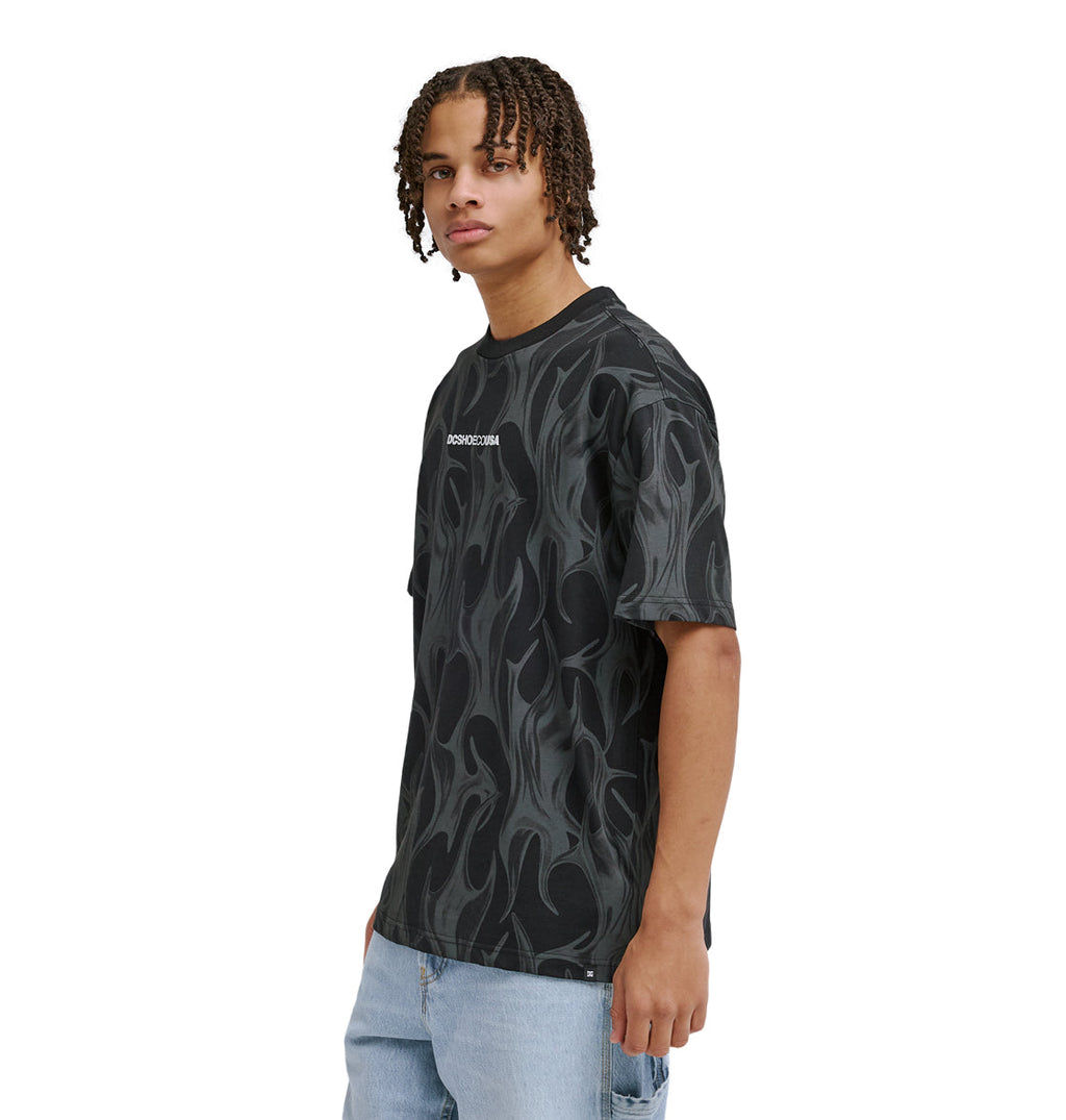 A young person with short dreadlocks wears the Reebok DC Liquid Fuego Hss black t-shirt with a gray flame pattern and light blue jeans, standing against a white background.