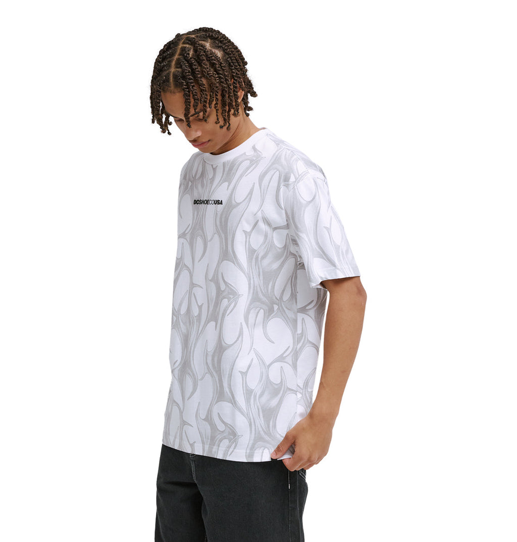 A person with braided hair wears the Reebok DC Liquid Fuego Hss white T-shirt featuring a subtle gray abstract flame pattern and small black chest text, paired with dark pants, looking down with one hand by their side.