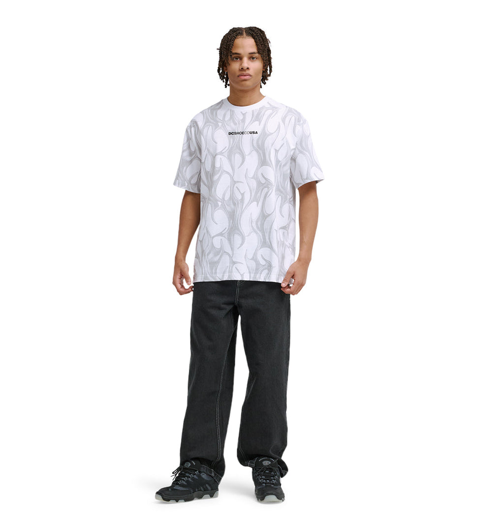A person with short, braided hair wears the Reebok DC Liquid Fuego Hss white patterned t-shirt with text on the chest, paired with black jeans and black sneakers, standing against a plain white background.