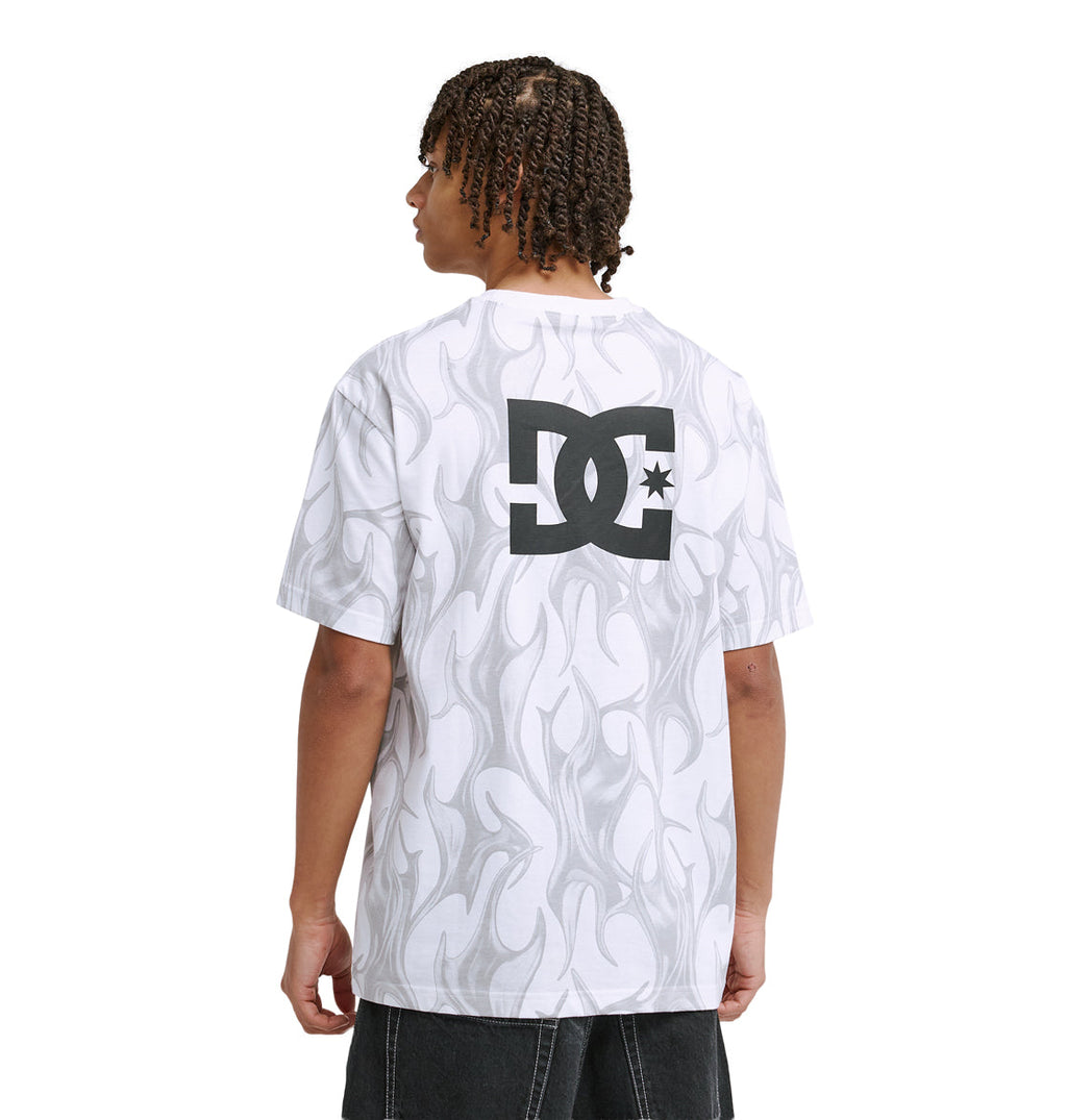 A person with short, curly hair wearing black pants and a white Reebok DC Liquid Fuego Hss t-shirt featuring gray flame patterns and a large black DC shoes logo on the back stands facing away from the camera.