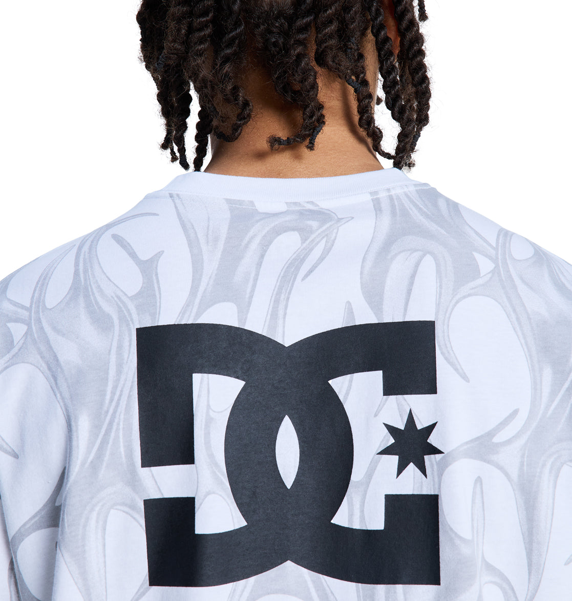 A person with short, twisted hair is seen from the back wearing the Reebok DC Liquid Fuego Hss, a white shirt featuring gray flame patterns and a bold black DC logo with a star printed on the upper back.