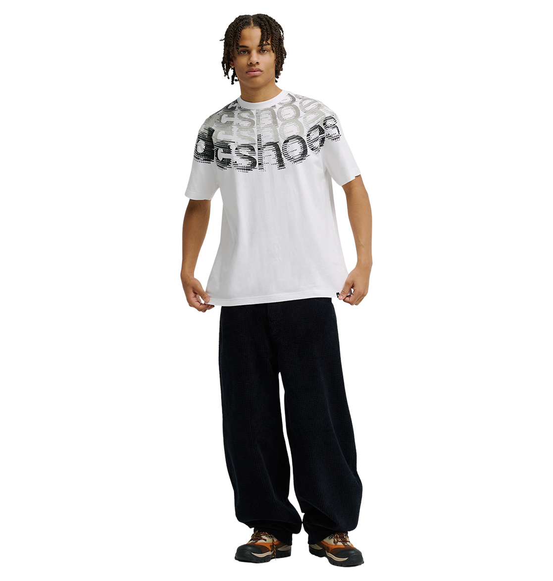 A person wearing the Men’s DC Omega 2 Tee from DC Shoes—a relaxed fit white graphic skate t-shirt with bold chest text—paired with loose black pants and brown sneakers, stands facing forward against a plain white background.