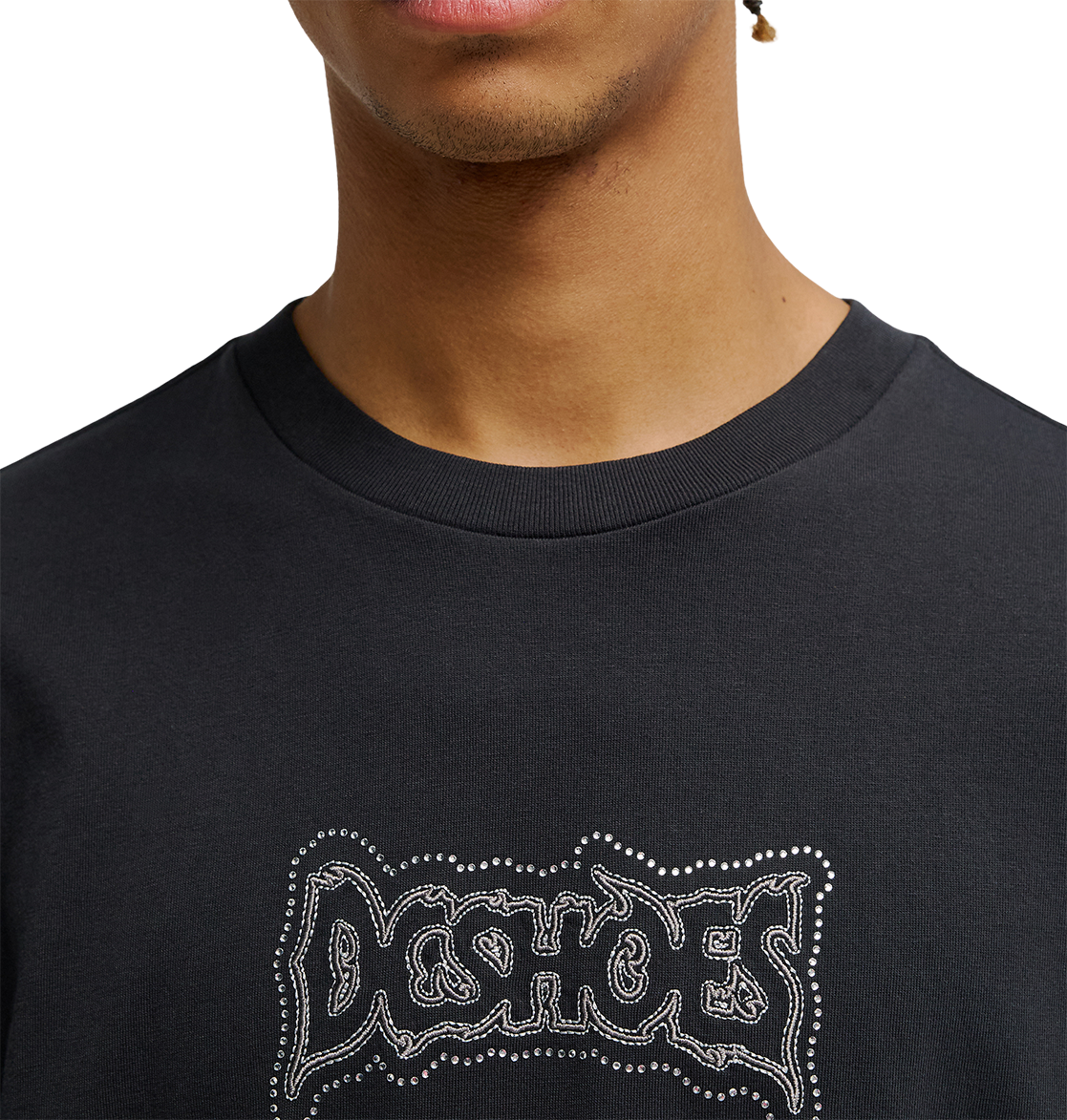 A person wearing the Men's DC Burning Diamonds Tee by DC Shoes, featuring a bold black graphic design with "DC SHOES" in a graffiti-style font accented by small white dots. Only the lower half of their face and upper torso are visible, emphasizing the streetwear aesthetic inspired by burning diamonds artwork.