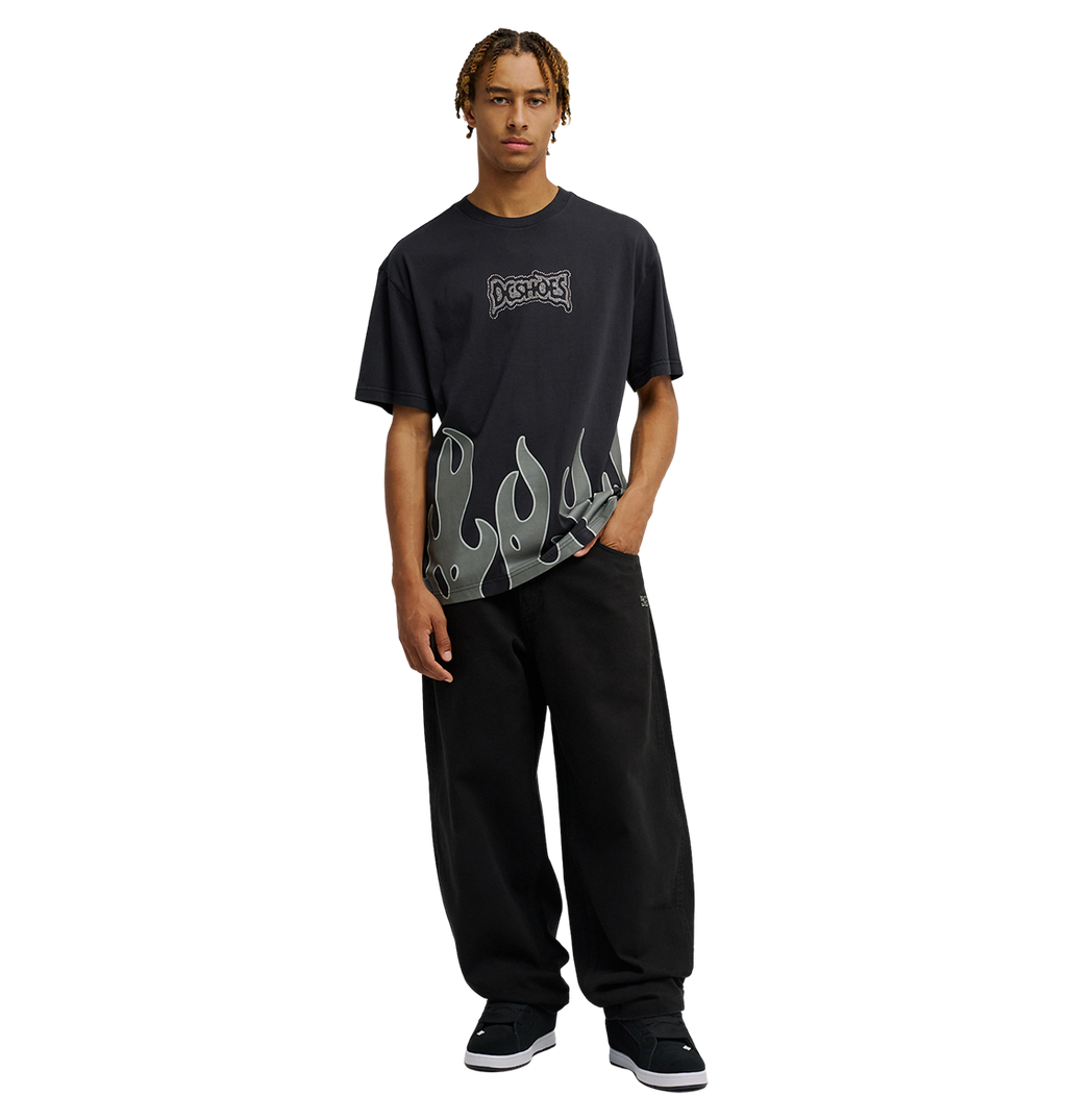 A person wearing the Men's DC Burning Diamonds Tee by DC Shoes, a black graphic skate shirt with a flame design along the bottom, and loose black pants stands facing forward against a plain white background.