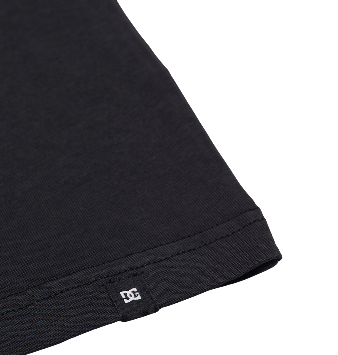 Close-up of the hem of the Men's DC Speratic Tee by DC Shoes in black, highlighting detailed stitching and a small white DC logo tag sewn into the bottom edge, set against a plain white background.