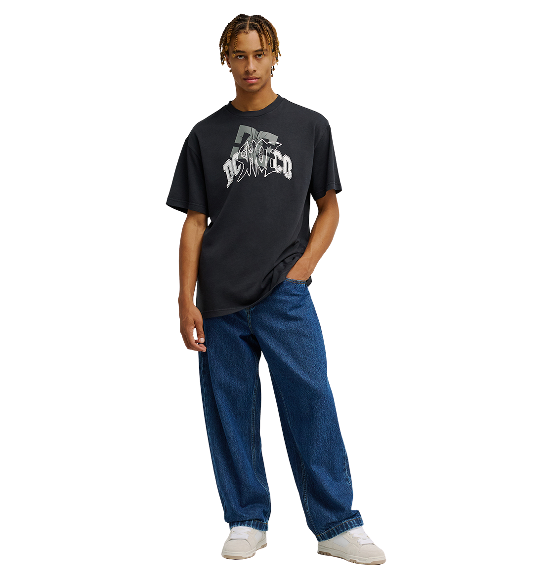 A person wearing the Men's DC Speratic Tee by DC Shoes—a black graphic skate shirt with heritage-inspired artwork—paired with loose blue jeans and white sneakers stands against a plain white background, one hand in their pocket.