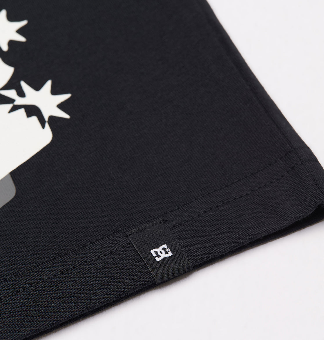Close-up of the hem on the Men's DC Side Flame Tee by DC Shoes, showcasing a small black fabric tag with a white DC Shoes logo, and part of the white graphic design above the hem—perfect for skate sessions.