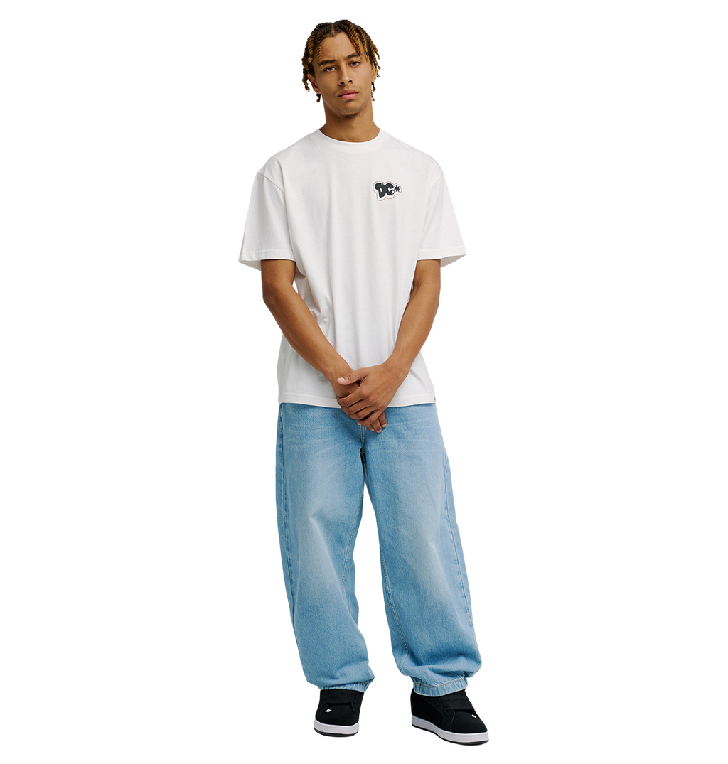 A person with short dreadlocks wears the Men's DC Side Flame Tee by DC Shoes—a relaxed fit white graphic skate tee—paired with light blue baggy jeans and black sneakers, standing against a plain white background with hands loosely clasped in front.