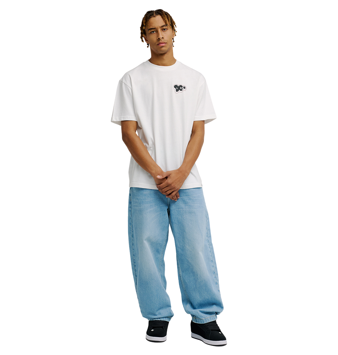 A person with short dreadlocks wears the Men's DC Side Flame Tee by DC Shoes—a relaxed fit white graphic skate tee—paired with light blue baggy jeans and black sneakers, standing against a plain white background with hands loosely clasped in front.