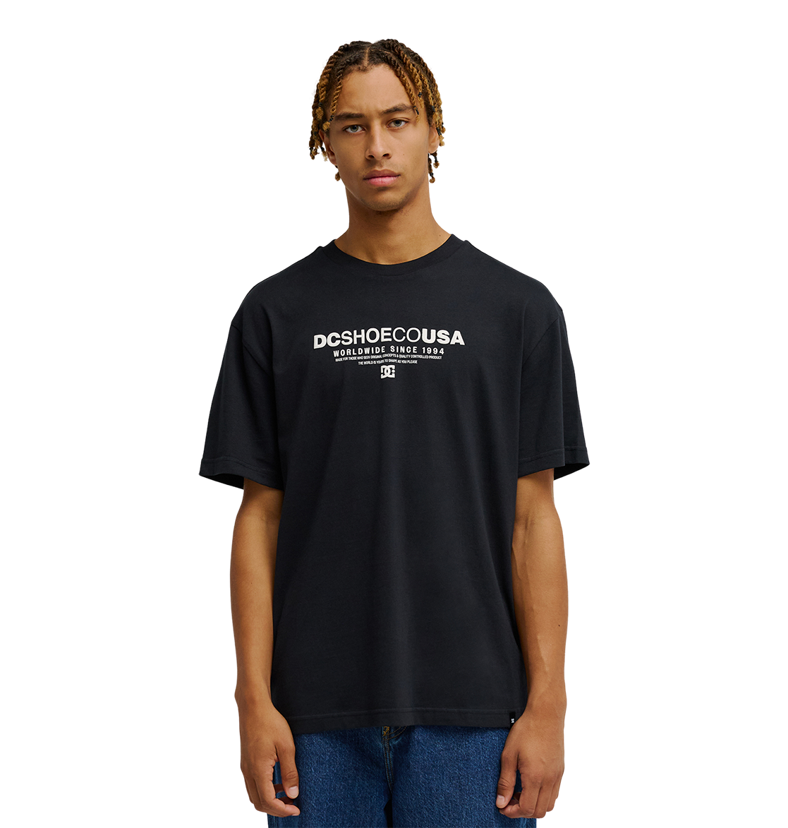 A person with short, braided hair wears the Men's DC Heritage Font Tee by DC Shoes in black, styled in a relaxed fit with blue jeans, standing against a plain white background.