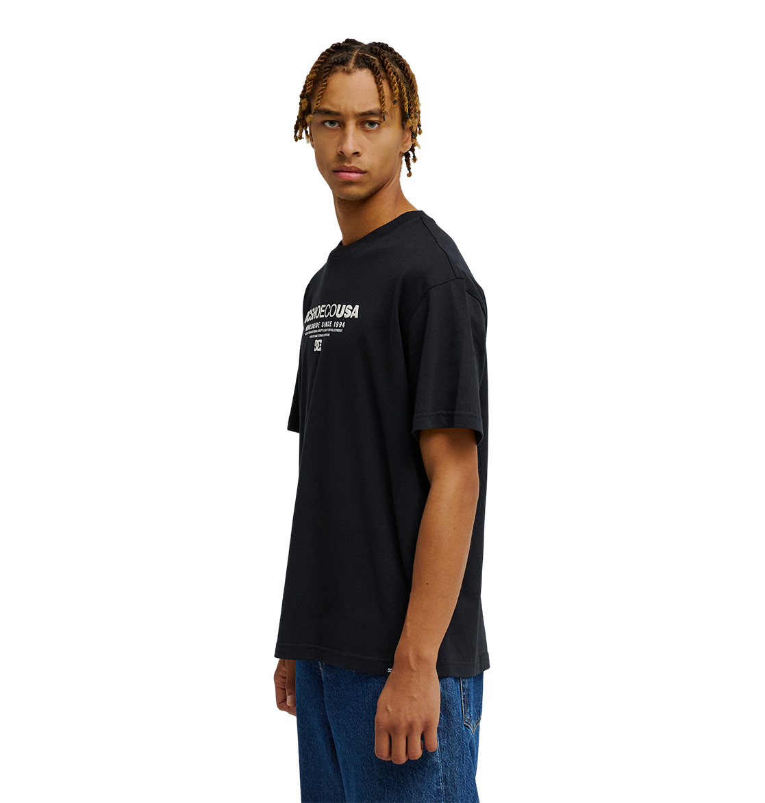 A person with short, braided hair wears the Men's DC Heritage Font Tee by DC Shoes—a relaxed fit graphic skate t-shirt with white heritage font—paired with blue jeans, standing sideways and looking forward against a plain white background.