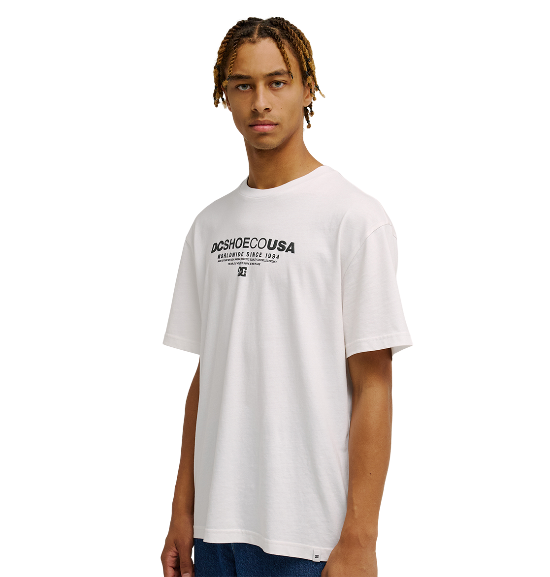 A young person with braided hair wears the Men's DC Heritage Font Tee by DC Shoes—a relaxed fit white graphic skate shirt—paired with blue jeans, standing against a plain white background.