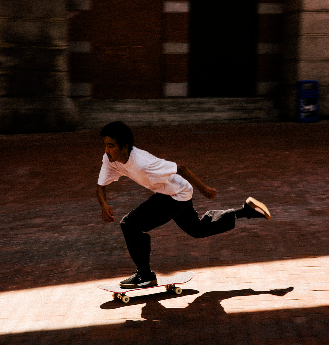 Wearing DC Shoes' Men's Fractal S Shoes, a skateboarder swiftly glides across a sunlit brick pavement, their clear shadow trailing behind—a perfect display of street-ready style and performance-driven comfort.