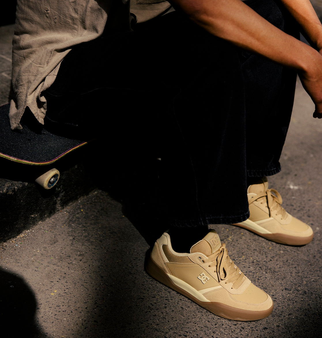 A person sits on a skateboard, wearing black pants and the DC Shoes Men's Fractal S Shoes in beige, as sunlight casts shadows on the ground—showcasing street-ready style and performance-driven comfort.