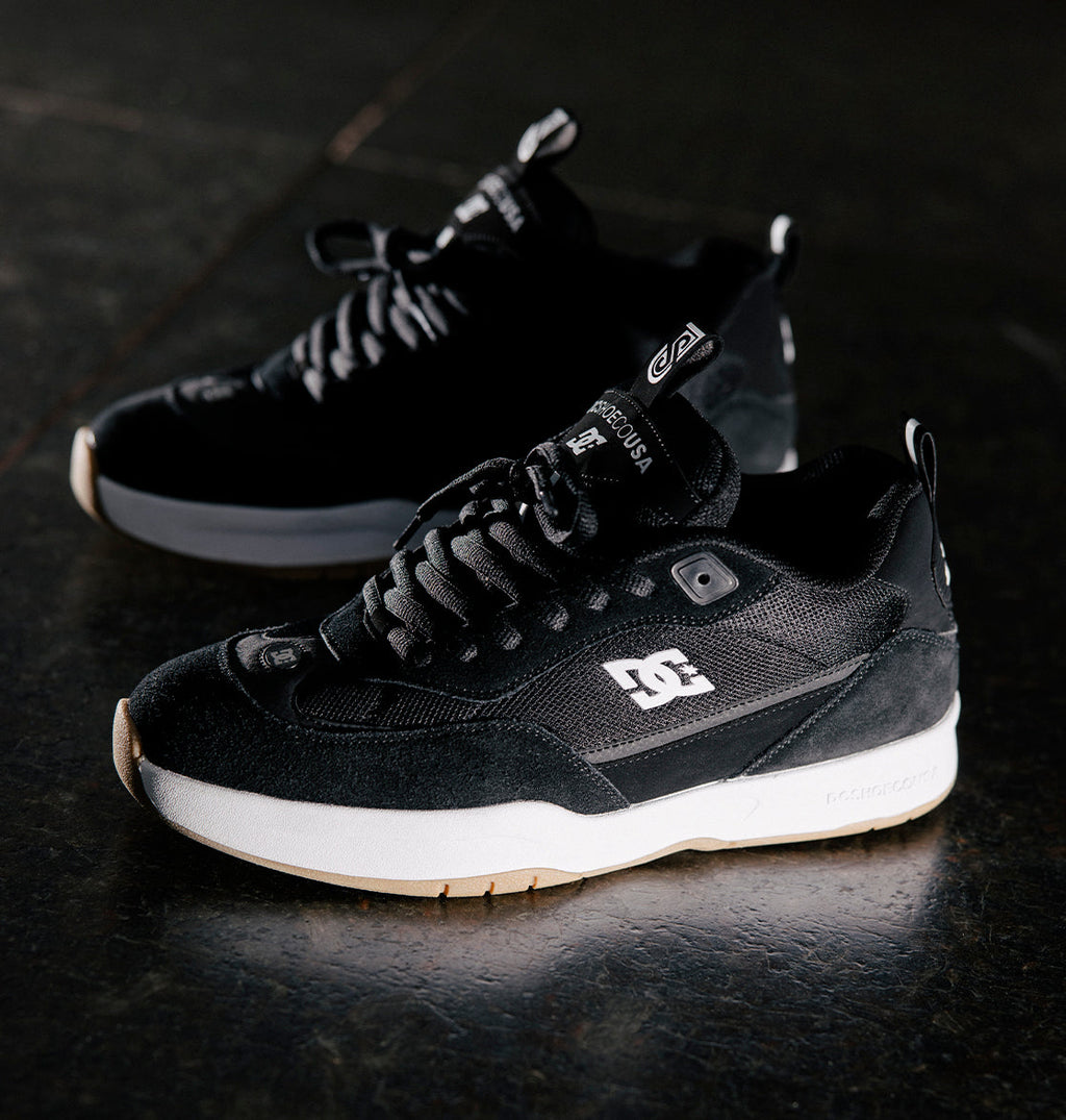 A pair of Men's JS-2 Shanahan Pro Skate Shoes by DC Shoes in black with white soles and branding, inspired by DC heritage, are displayed on a dark surface with one shoe slightly ahead of the other.